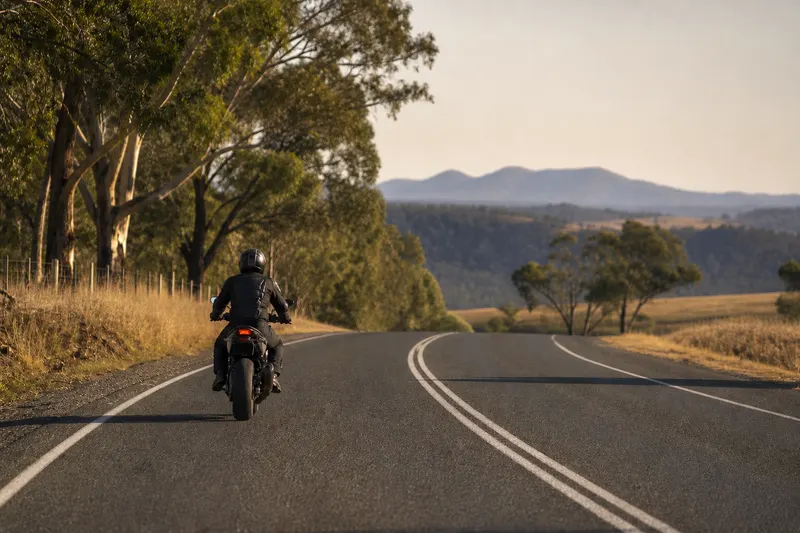 A rider in full gear seen from behind, riding away along a two lane country road with gum trees on the verges.